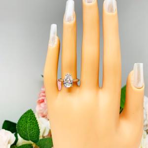 Mannequin hand with long white shimmery nails, displayed against a soft white-gray background with pink and white floral accents. The hand is wearing an elegant 4-prong sterling silver cathedral engagement ring featuring a 12x8mm precision-cut pear cubic zirconia.