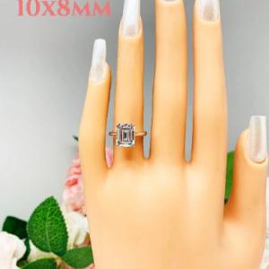 Mannequin hand with long white shimmery nails, displayed against a soft white-gray background with pink and white floral accents. The hand is wearing a sterling silver solitaire cathedral engagement ring set with a 10x8mm emerald cut cubic zirconia.