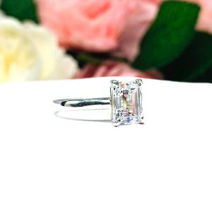 Sterling silver solitaire cathedral engagement ring set with a 10x8mm emerald cut cubic zirconia. The ring is resting on a white surface with a background of pink and white flowers.