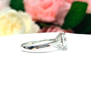Sterling silver solitaire cathedral engagement ring set with a 10x8mm emerald cut cubic zirconia. The ring is resting on a white surface with a background of pink and white flowers.