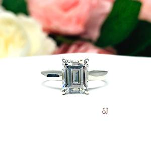 Sterling silver solitaire cathedral engagement ring set with a 10x8mm emerald cut cubic zirconia. The ring is resting on a white surface with a background of pink and white flowers.