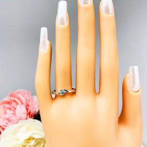 A display hand with long white fingernails standing up and with the top of the hand facing forward. The display hand is wearing a sterling silver ring with a bypass design and 3mm round blue topaz set in it on the ring finger.