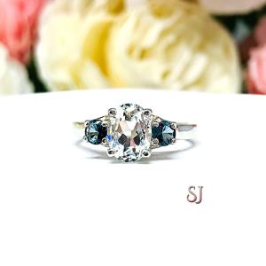 A sterling silver 3-stone ring with a 9x7mm oval-shaped white topaz and two smaller round medium tone London blue topaz gemstones on either side of the center stone. The ring is on a white surface with a background of pastel flowers.