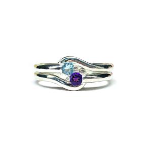Two stacked silver round gemstone bypass rings with thin bands. The top ring has a 3mm round swiss blue topaz gemstone and fits perfectly with the bottom ring, a 3mm round purple amethyst gemstone. The gems sit an an angle and are touching. The modern setting design curves around each gemstone. The rings are set against a white background.