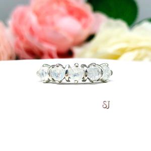 A sterling silver ring featuring five 4mm round natural rainbow moonstones in a row. The ring has rounded bars separating each stone. Displayed on a white surface with pastel floral background.