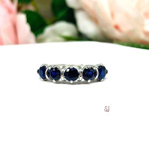 A sterling silver ring featuring five 4mm round lab created blue sapphires in a row. The ring has rounded bars separating each stone. Displayed on a white surface with pastel floral background.