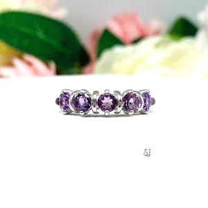 A sterling silver ring featuring five 4mm round medium purple tone amethysts in a row. The ring has rounded bars separating each stone. Displayed on a white surface with pastel floral background.