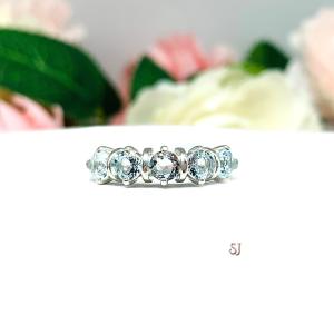 A sterling silver ring featuring five 4mm round sky blue topaz gemstones in a row. The ring has rounded bars separating each stone. Displayed on a white surface with pastel floral background.