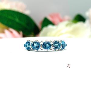 A sterling silver ring featuring five 4mm round swiss blue topaz gemstones in a row. The ring has rounded bars separating each stone. Displayed on a white surface with pastel floral background.