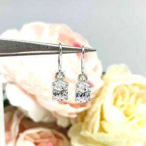 A pair of silver dangle drop basket style earrings set with brilliant cut oval cubic zirconia. The cz dangles are hanging from ear wires and set against a background of soft-focus pink and white flowers.
