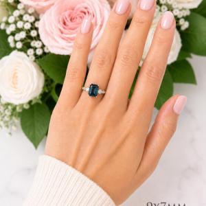 A woman's hand wearing a sterling silver ring featuring a 9x7mm emerald cut London blue topaz flanked by a round moissanite on each side. Model image for size and styling reference — not a photo of the actual ring.