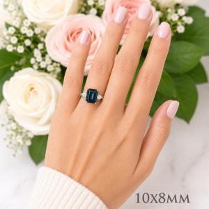 A woman's hand wearing a sterling silver ring featuring a 10x8mm emerald cut London blue topaz flanked by a round moissanite on each side. Model image for size and styling reference — not a photo of the actual ring.