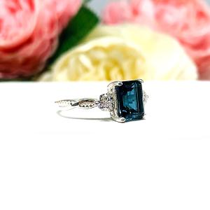Close-up of an 8x6mm emerald-cut London Blue Topaz ring with moissanite side stones, set in a sterling silver twist band with milgrain detailing; soft floral background enhances the elegance.