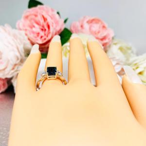 Mannequin hand with long white shimmery nails, displayed against a soft white-gray background with pink and white floral accents. The hand is wearing an 8x6mm emerald-cut London Blue Topaz ring with moissanite side stones, set in a sterling silver twist band with milgrain detailing.
