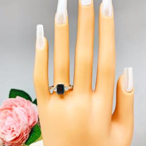 Mannequin hand with long white shimmery nails, displayed against a soft white-gray background with pink and white floral accents. The hand is wearing an 8x6mm emerald-cut London Blue Topaz ring with moissanite side stones, set in a sterling silver twist band with milgrain detailing.
