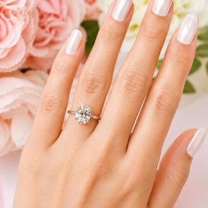 A woman's hand wearing a sterling silver 6-prong solitaire engagement ring set with a 10x8mm near colorless oval cubic zirconia. It is resting on a white surface with soft-focus pink and white flowers in the background. Model image for size and styling reference — not a photo of the actual ring.