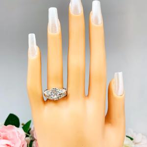 Mannequin hand with long white shimmery nails, displayed against a soft white-gray background with pink and white floral accents. The hand is wearing a sterling silver 5-stone curved wedding band featuring 3mm round precision faceted moissanite stones in prong settings. The ring is paired with a round with pear side stone 3-stone moissanite engagement ring. This ring is not included.