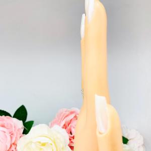 Mannequin hand with long white shimmery nails, displayed against a soft white-gray background with pink and white floral accents. The hand is wearing a sterling silver stackable 5-stone wedding band featuring 3mm round authentic moissanite stones in prong settings.