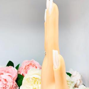 Mannequin hand with long white shimmery nails, displayed against a soft white-gray background with pink and white floral accents. The hand is wearing a sterling silver 5-stone curved wedding band featuring 3mm round precision faceted near colorless cz stones in prong settings.