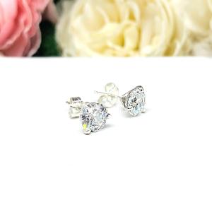 A pair of sterling silver 3-prong martini stud earrings with 6.5mm colorless old European cut cubic zirconias. The earrings are resting on a white surface with a background of pink and white flowers.