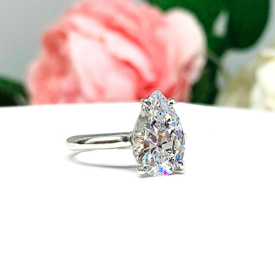 Elegant 4-prong sterling silver cathedral engagement ring set featuring a 12x8mm precision-cut pear cubic zirconia.  Displayed on a white surface with soft pastel floral background.