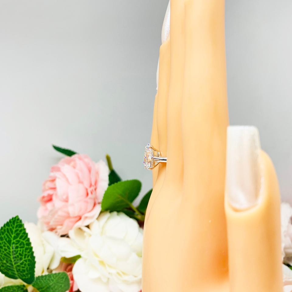 Mannequin hand with long white shimmery nails, displayed against a soft white-gray background with pink and white floral accents. The hand is wearing an elegant 4-prong sterling silver cathedral engagement ring featuring a 12x8mm precision-cut pear cubic zirconia.