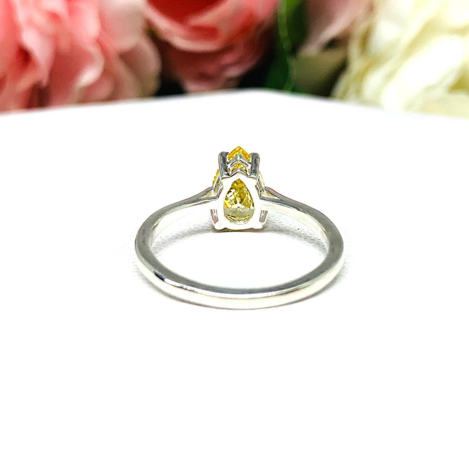 A sterling silver engagement ring set with a 9x6mm pear-cut canary yellow cz. Displayed on a white surface with soft pastel floral background.