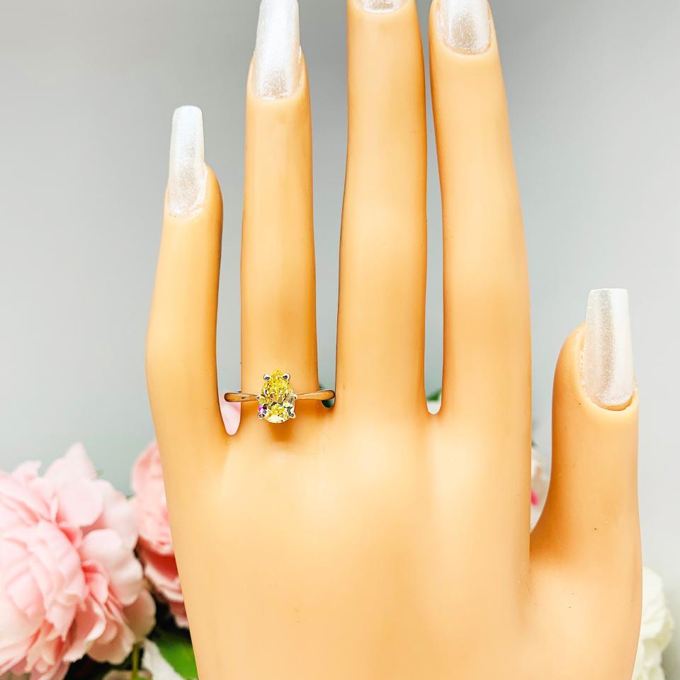 Mannequin hand with long white shimmery nails, displayed against a soft white-gray background with pink and white floral accents. The hand is wearing a sterling silver engagement ring set with a 9x6mm pear-cut canary yellow cz.