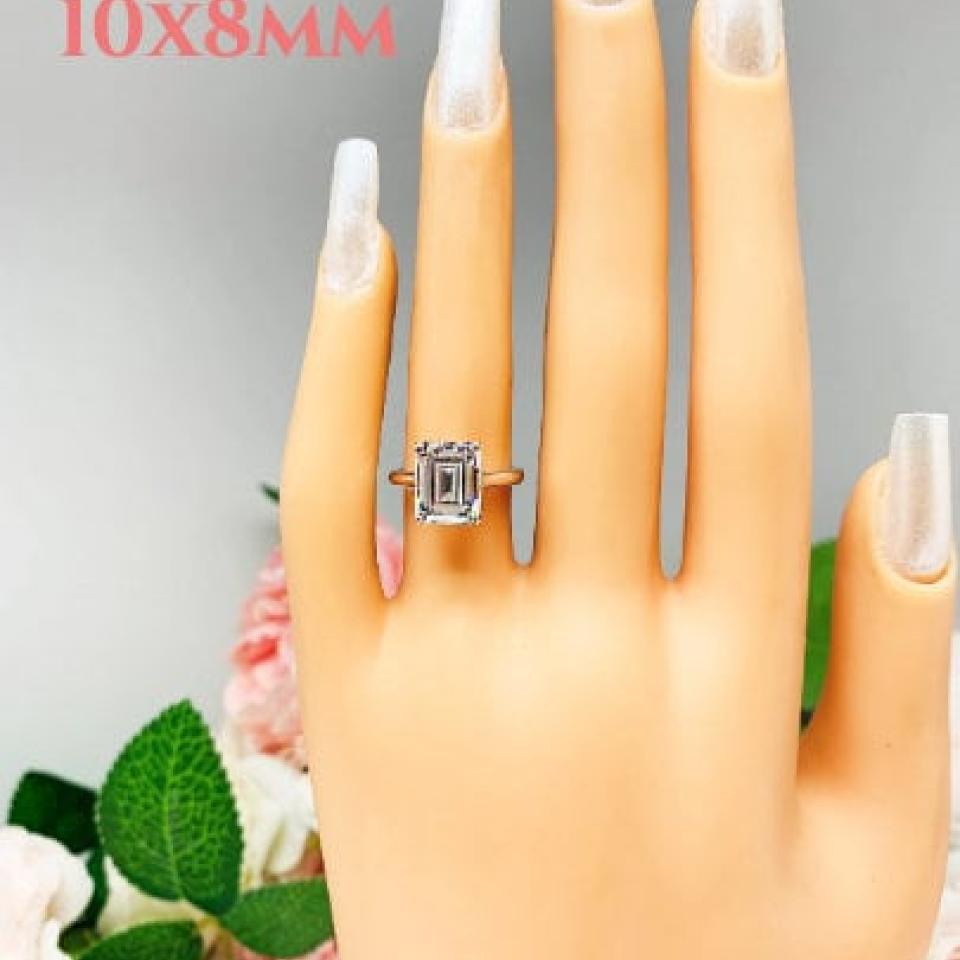 Mannequin hand with long white shimmery nails, displayed against a soft white-gray background with pink and white floral accents. The hand is wearing a sterling silver solitaire cathedral engagement ring set with a 10x8mm emerald cut cubic zirconia.