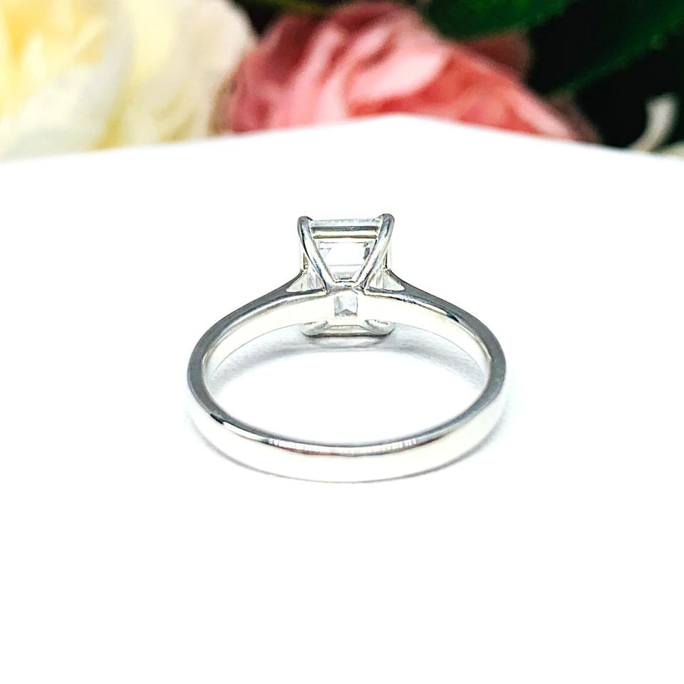 Sterling silver solitaire cathedral engagement ring set with a 10x8mm emerald cut cubic zirconia. The ring is resting on a white surface with a background of pink and white flowers.