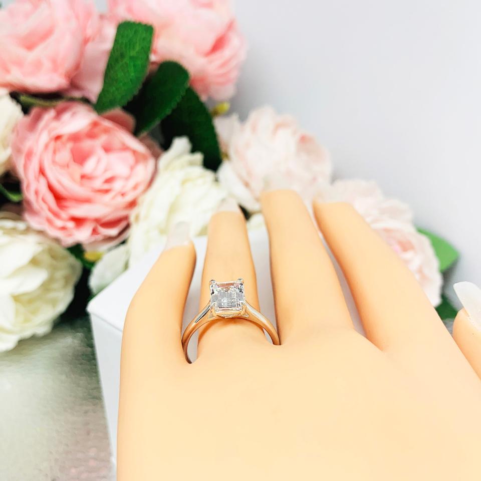 Mannequin hand with long white shimmery nails, displayed against a soft white-gray background with pink and white floral accents. The hand is wearing a sterling silver solitaire cathedral engagement ring set with a 10x8mm emerald cut cubic zirconia.