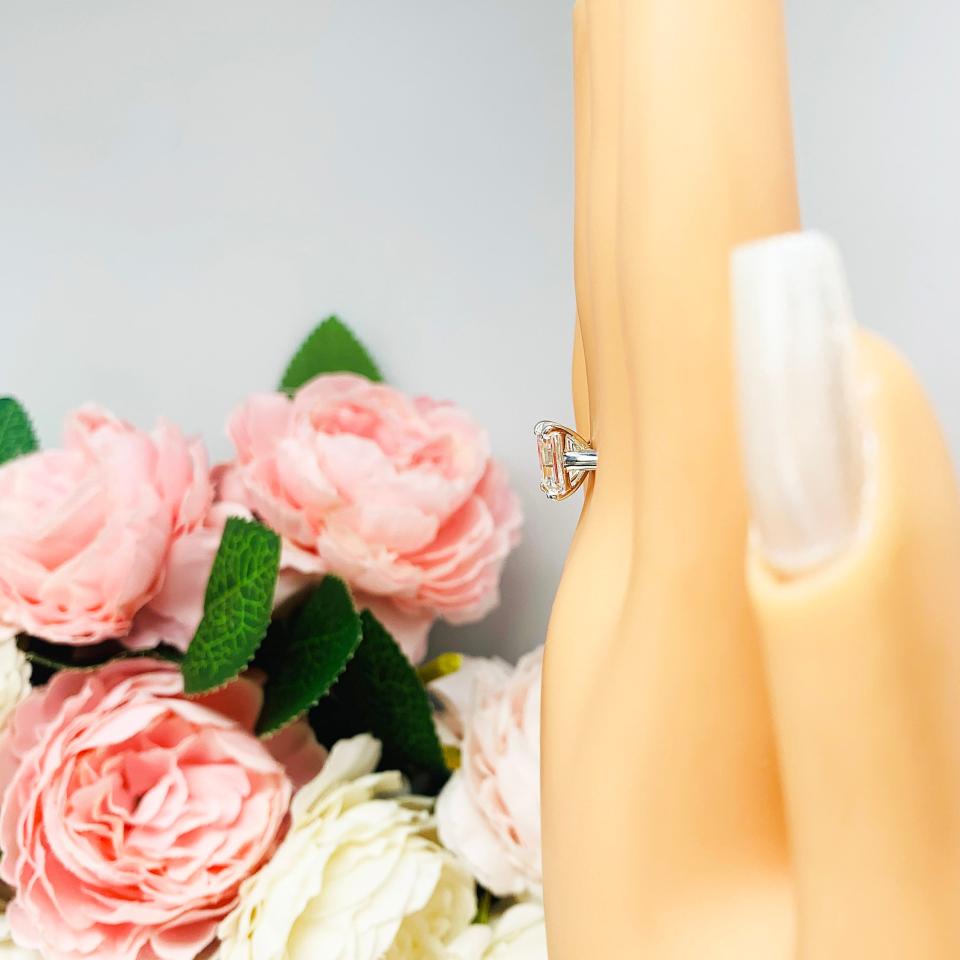 Mannequin hand with long white shimmery nails, displayed against a soft white-gray background with pink and white floral accents. The hand is wearing a sterling silver solitaire cathedral engagement ring set with a 10x8mm emerald cut cubic zirconia.