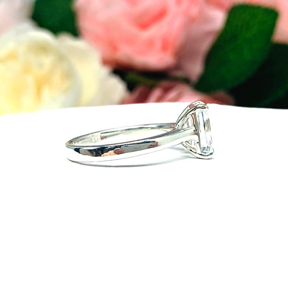 Sterling silver solitaire cathedral engagement ring set with a 10x8mm emerald cut cubic zirconia. The ring is resting on a white surface with a background of pink and white flowers.