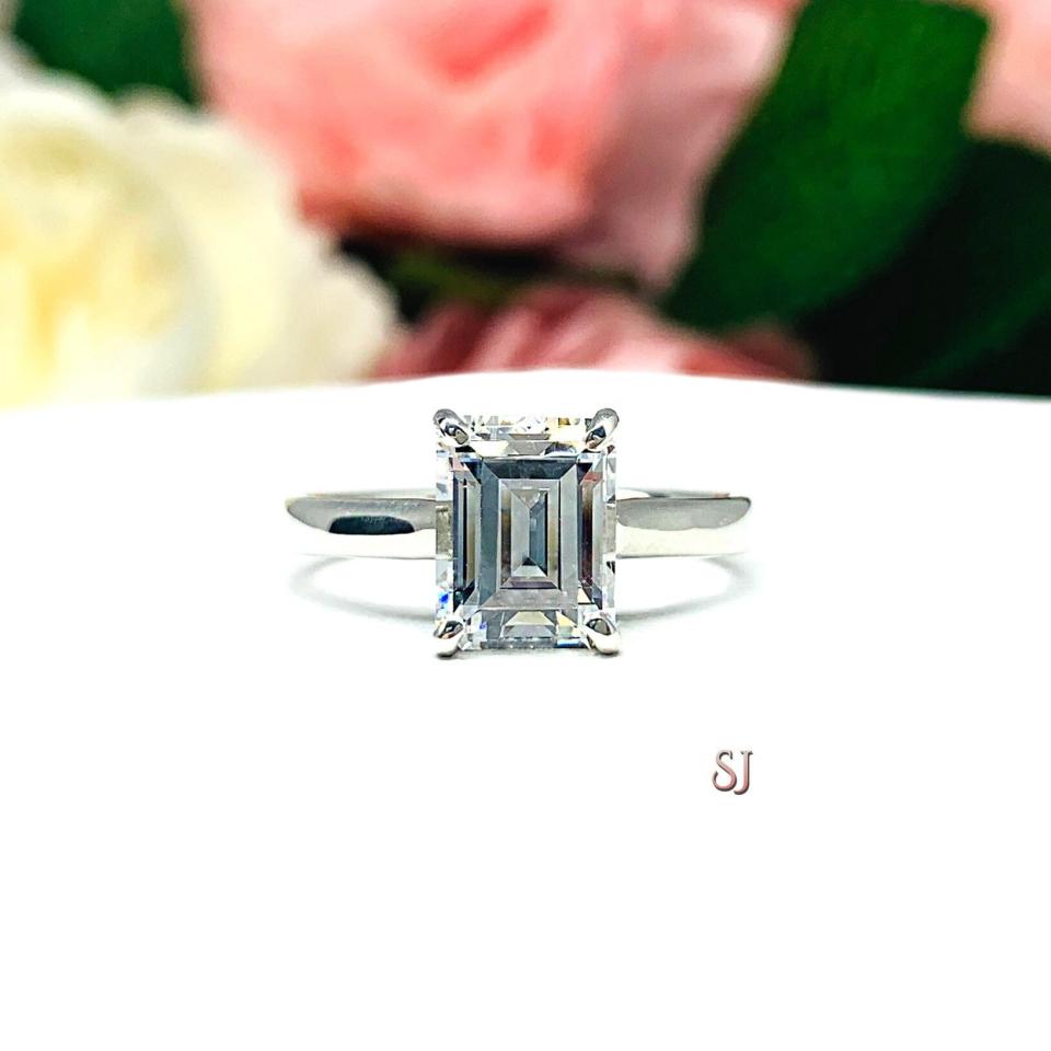Sterling silver solitaire cathedral engagement ring set with a 10x8mm emerald cut cubic zirconia. The ring is resting on a white surface with a background of pink and white flowers.