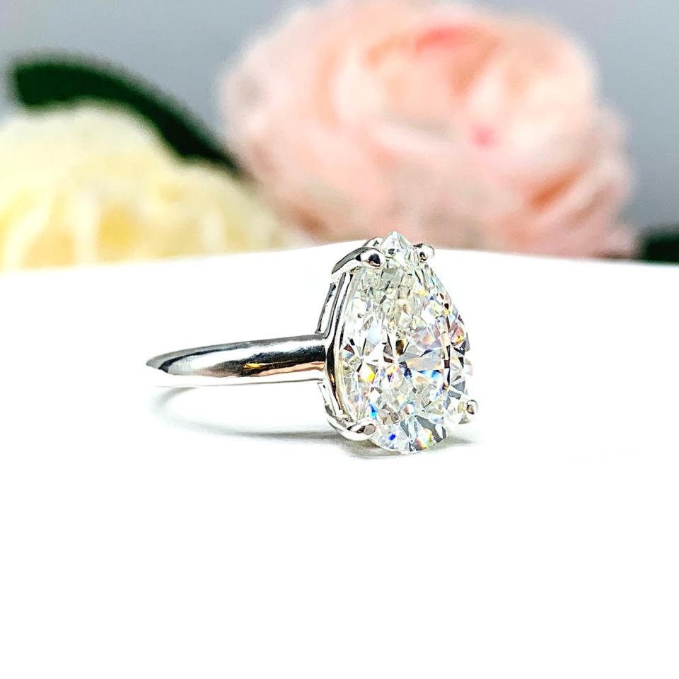 An elegant 4-prong sterling silver engagement ring with a 3mm wide band featuring a 13x9mm near colorless precision-cut pear cubic zirconia. Displayed on a white surface with a floral background.