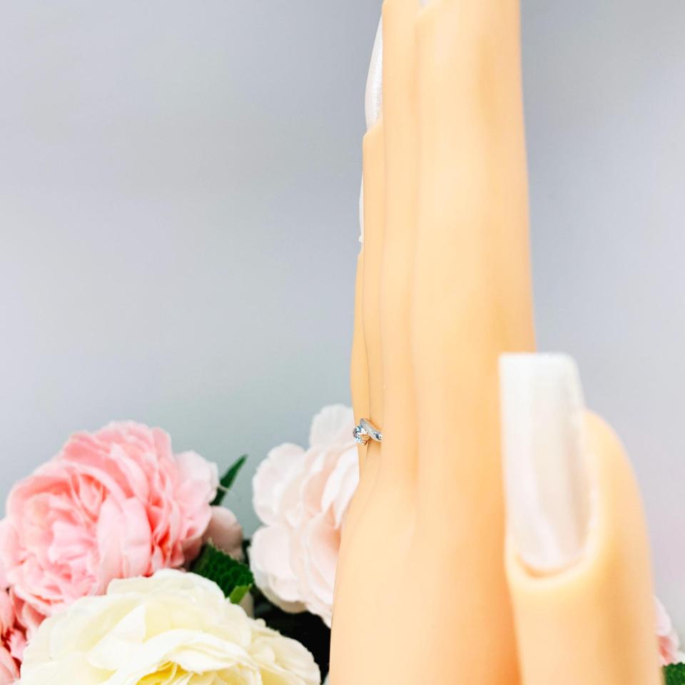 A display hand with long white fingernails standing up and with the top of the hand facing toward the left to show the side view. The display hand is wearing a sterling silver ring with a bypass design and 3mm round sky blue topaz gemstone set in it on the ring finger.