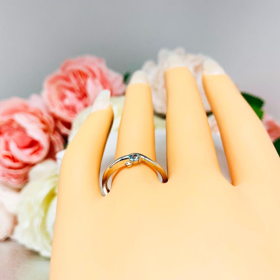 A display hand with long white fingernails laying down with the top of the hand facing up and fingernails facing toward the back. The display hand is wearing a sterling silver ring with a bypass design and 3mm round light blue topaz gemstone set in it on the ring finger.