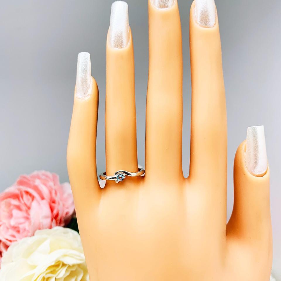 A display hand with long white fingernails standing up and with the top of the hand facing forward. The display hand is wearing a sterling silver ring with a bypass design and 3mm round blue topaz set in it on the ring finger.