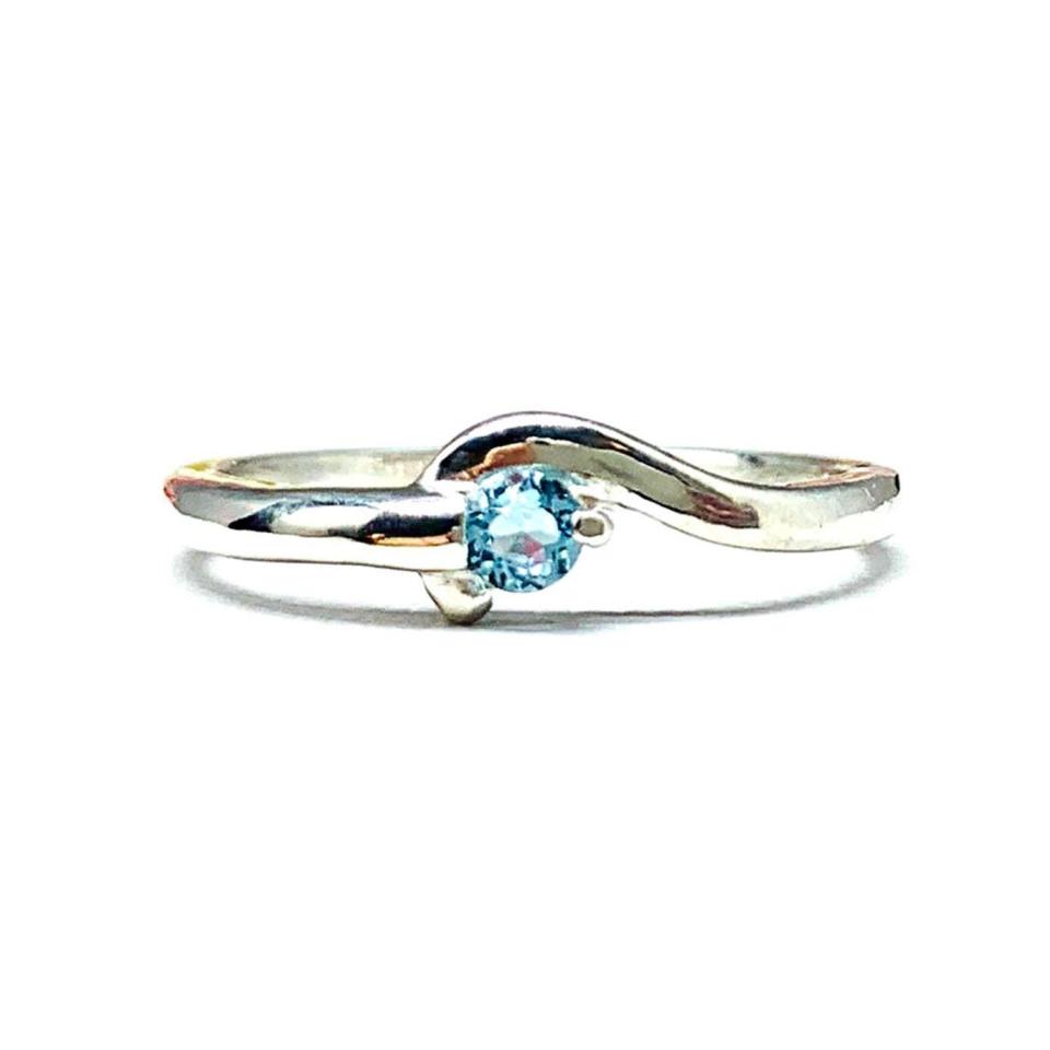 A sterling silver round gemstone bypass ring with a thin band. The ring is set with 3mm round swiss blue topaz gemstone. The modern setting design curves around the gemstone. The ring is facing toward the back and is set against a white background.