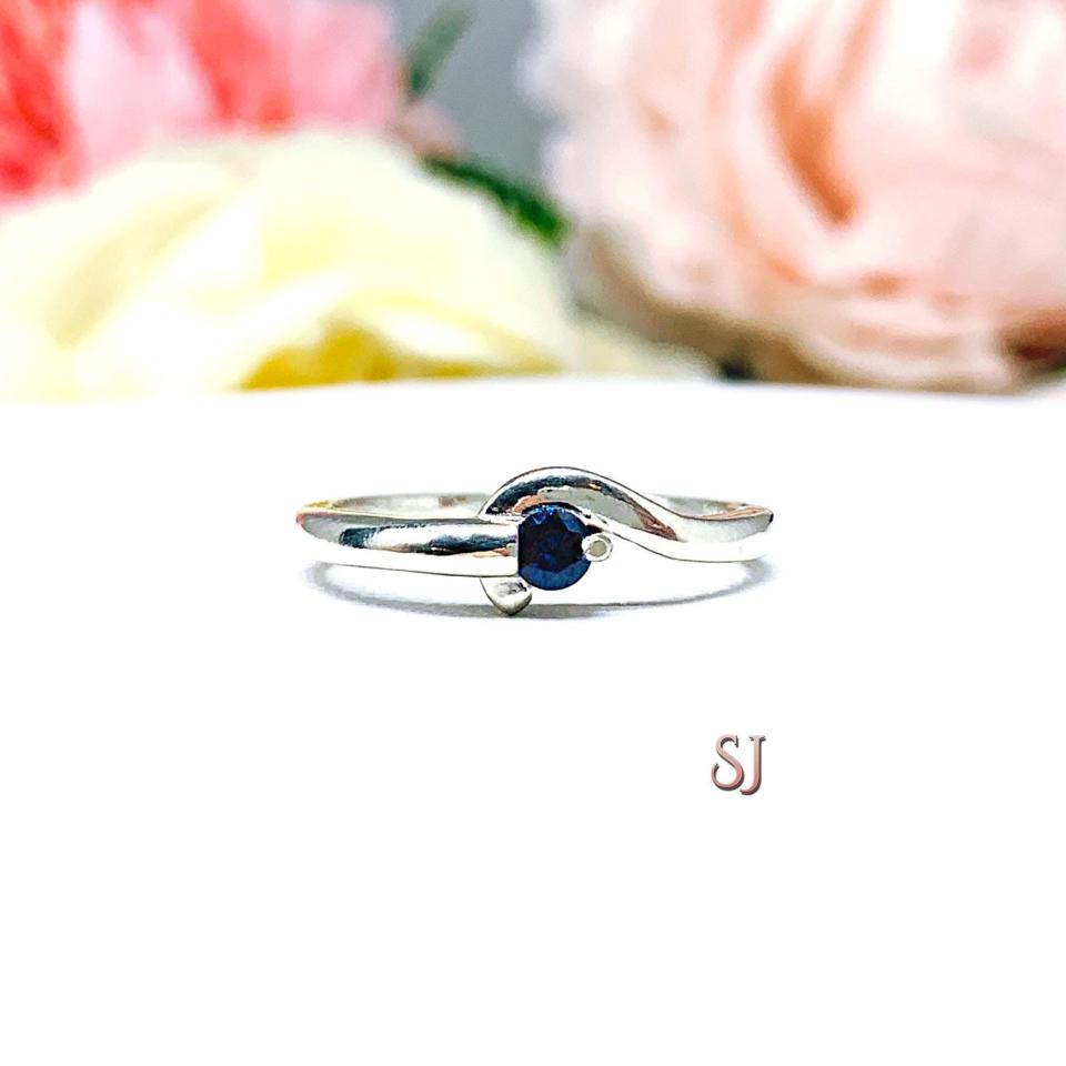 A sterling silver round gemstone bypass ring with a thin band. The ring is set with 3mm round lab blue sapphire. The modern setting design curves around the gemstone. The ring is facing toward the back and is set against a white background.