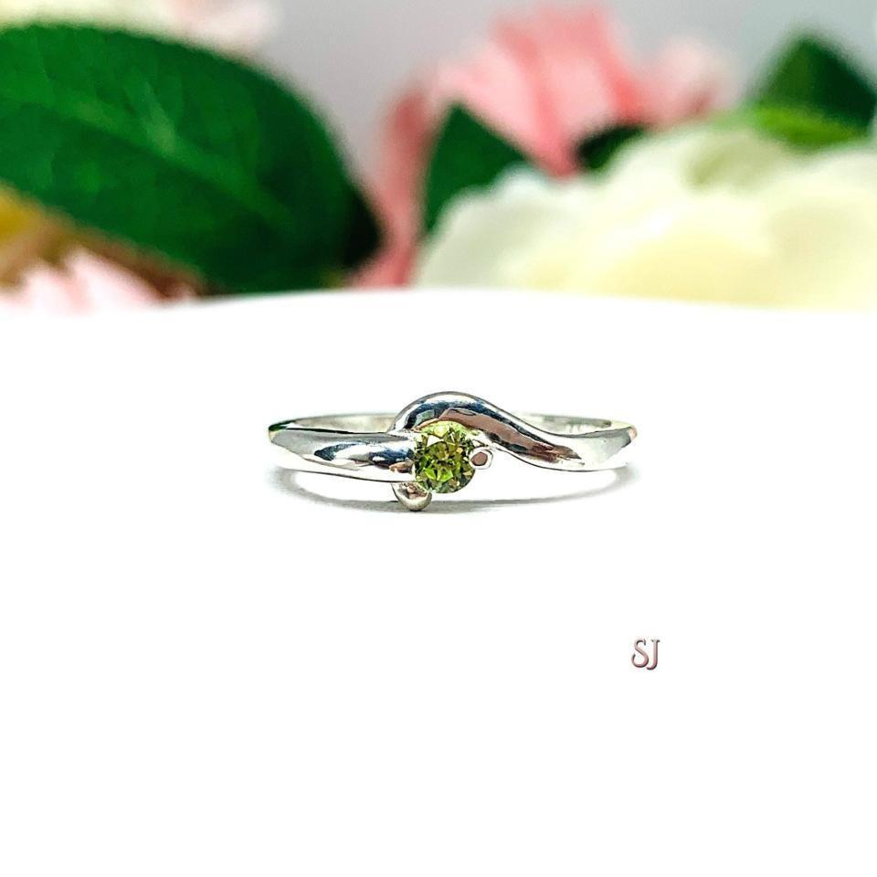 A silver round gemstone bypass ring with a thin band. The ring is set with a 3mm round peridot gemstone. The modern setting design curves around the gemstone. The ring is facing toward the back and is set against a white background.