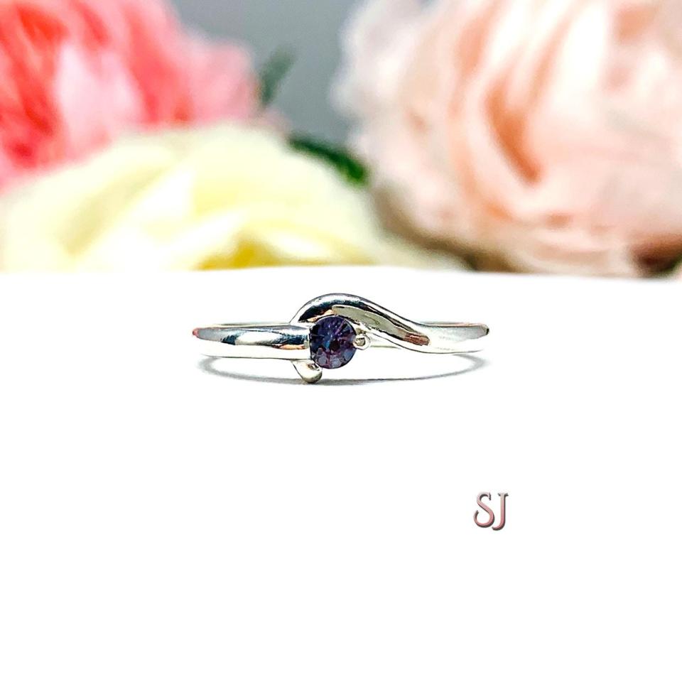A sterling silver round gemstone bypass ring with a thin band. The ring is set with 3mm round lab alexandrite. The modern setting design curves around the gemstone. The ring is facing toward the back and is set against a white background.
