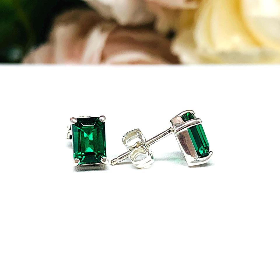 May include: Emerald-cut stud earrings with a vibrant green gemstone. The earrings are set in a silver-colored metal, featuring a rectangular emerald stone. The stud posts are visible, and the earrings are displayed on a white surface with a soft-focus background.