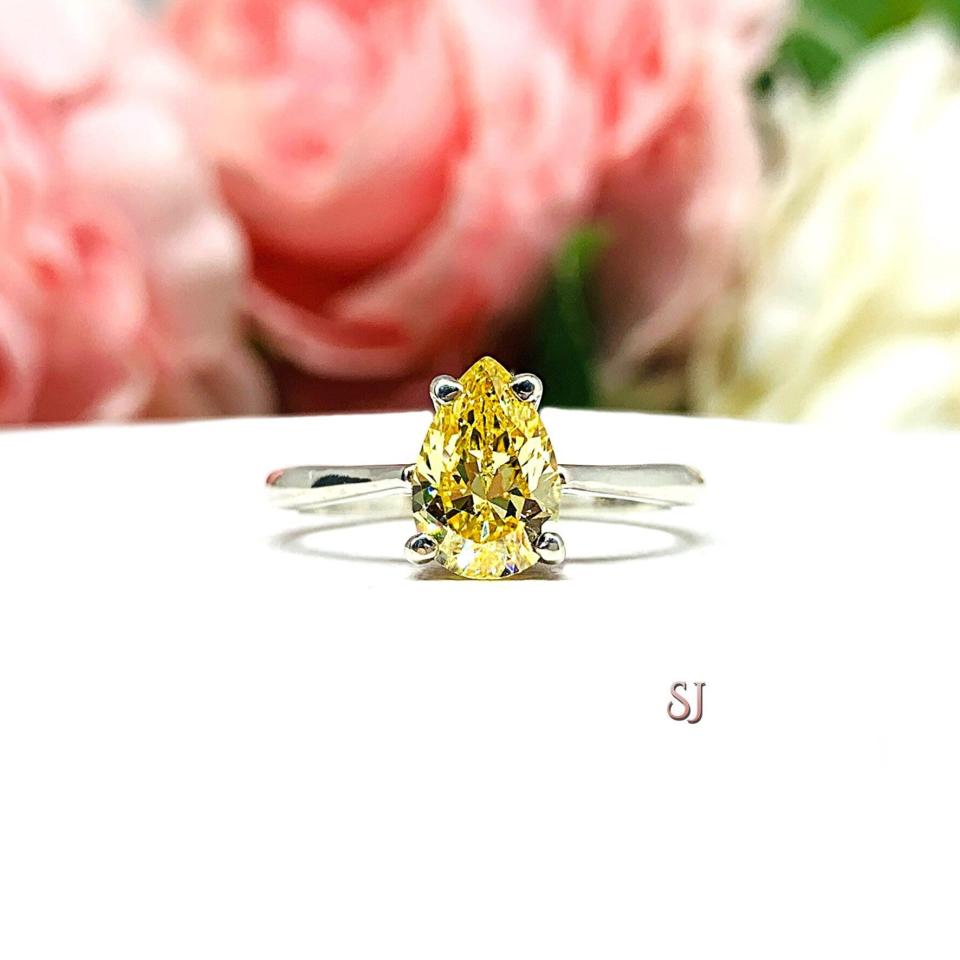 A sterling silver engagement ring set with a 9x6mm pear-cut canary yellow cz. Displayed on a white surface with soft pastel floral background.
