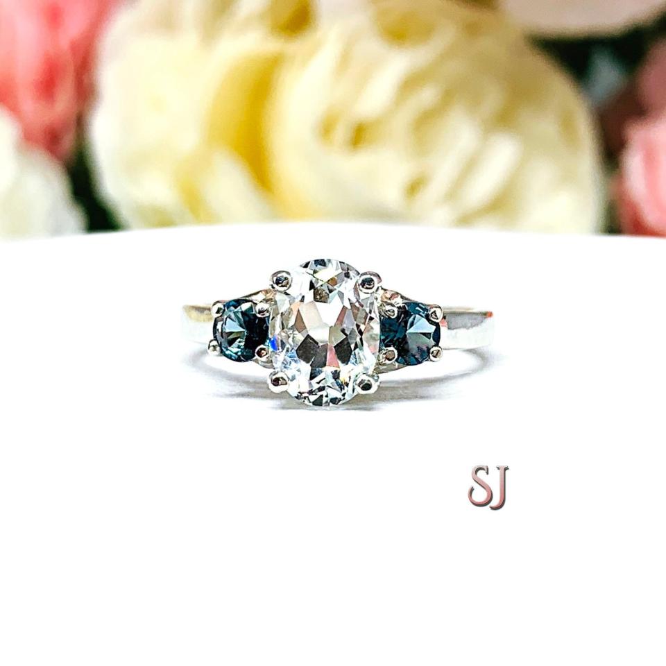 A sterling silver 3-stone ring with a 9x7mm oval-shaped white topaz and two smaller round medium tone London blue topaz gemstones on either side of the center stone. The ring is on a white surface with a background of pastel flowers.