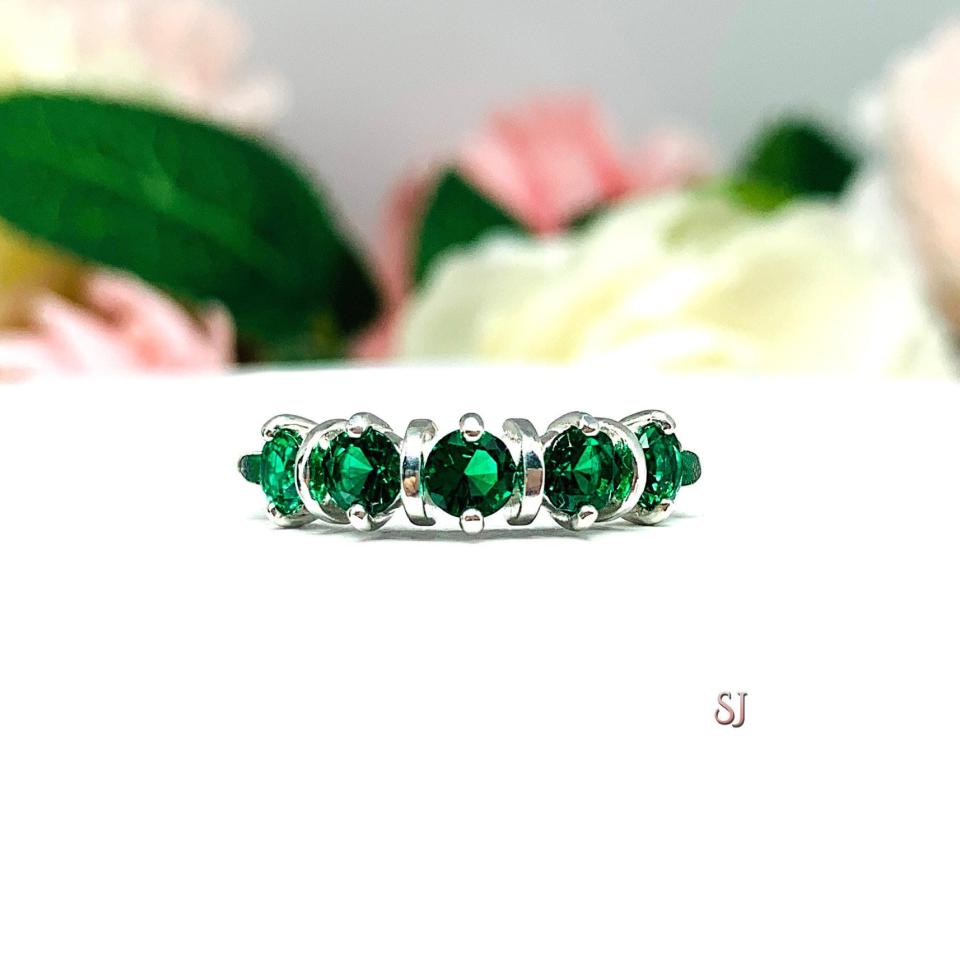 A sterling silver ring featuring five 4mm round lab created emeralds in a row. The ring has rounded bars separating each stone. Displayed on a white surface with pastel floral background.