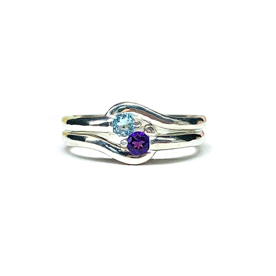 Two stacked silver round gemstone bypass rings with thin bands. The top ring has a 3mm round swiss blue topaz gemstone and fits perfectly with the bottom ring, a 3mm round purple amethyst gemstone. The gems sit an an angle and are touching. The modern setting design curves around each gemstone. The rings are set against a white background.
