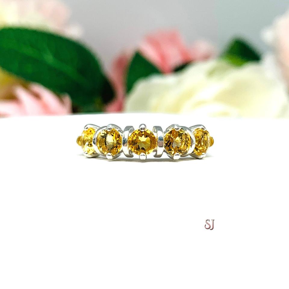 A sterling silver ring featuring five 4mm round golden citrine in a row. The ring has rounded bars separating each stone. Displayed on a white surface with pastel floral background.