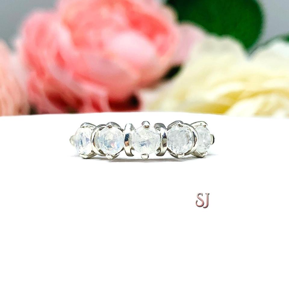 A sterling silver ring featuring five 4mm round natural rainbow moonstones in a row. The ring has rounded bars separating each stone. Displayed on a white surface with pastel floral background.