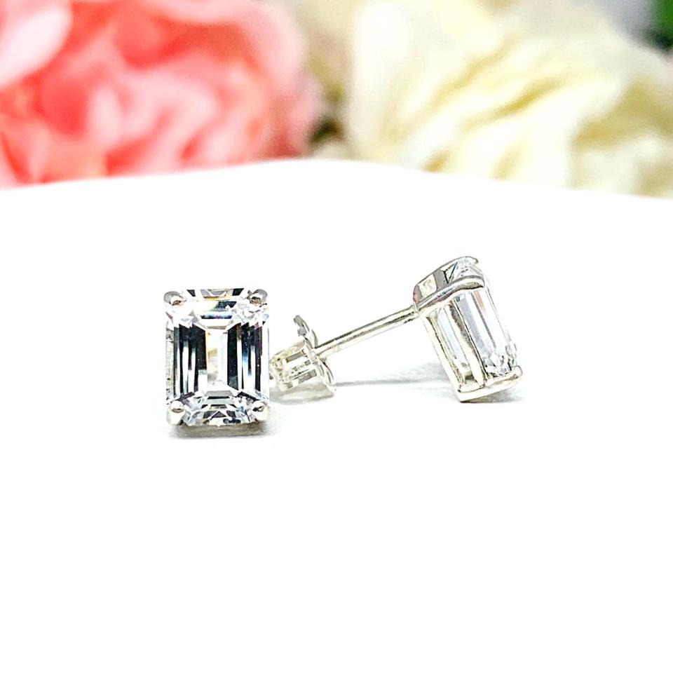 A pair of silver earrings with clear emerald cut lab created white sapphire stones. The earrings are on a white surface. The left earring is facing forward to showcase the crisp line faceting, and the right earring is facing the right to display the post at the top and butterfly friction clasp.